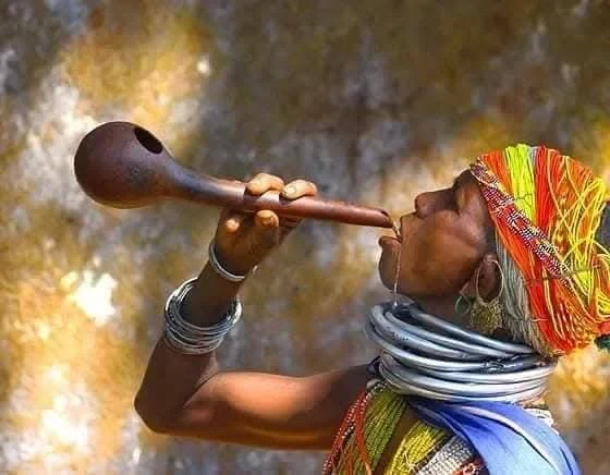 A tribal woman in bead necklaces and heavy metal neck rings playing a long wind instrument made from a gourd