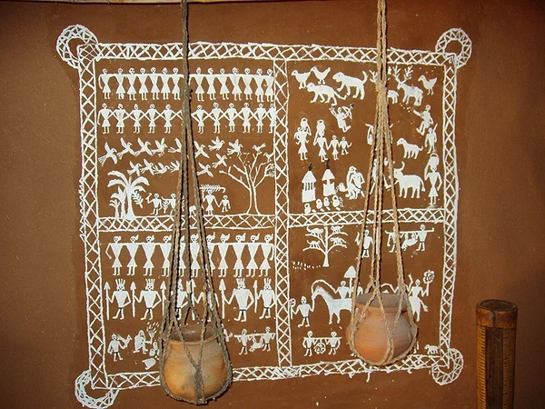 Saura idital wall painting in white on a terracotta mud wall, showing rows of figures, animals and a tree inside a woven border, with two clay pots hanging in front