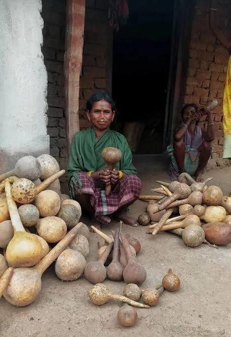 A woman seated on the ground behind heaps of dried bottle gourds of many shapes, outside a village house